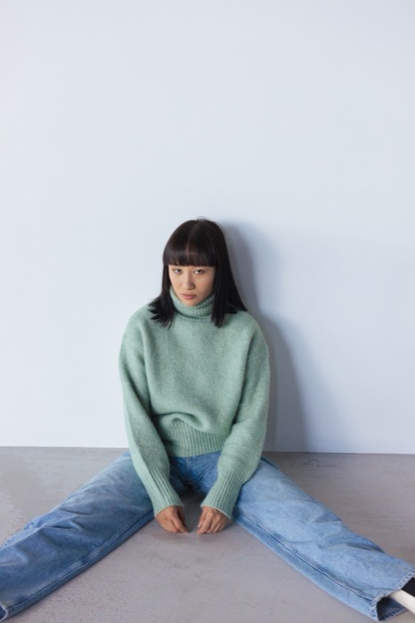 Sage jumper and wide jeans, seated against a white wall